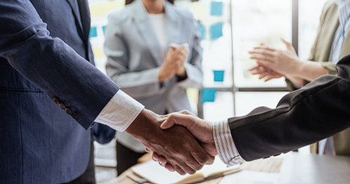 Two colleagues shaking hands in an office while others applaud