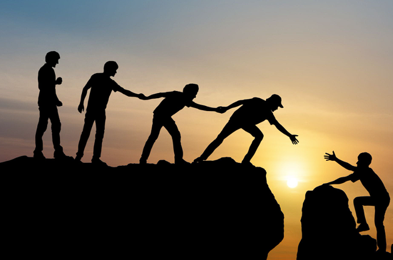 Silhouettes of people on a cliff at sunset, reaching down to help one person climb up.